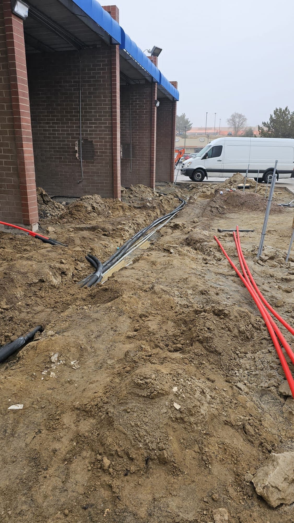 Open trench beside a brick storefront with new pipe laid in it and a white work van parked nearby